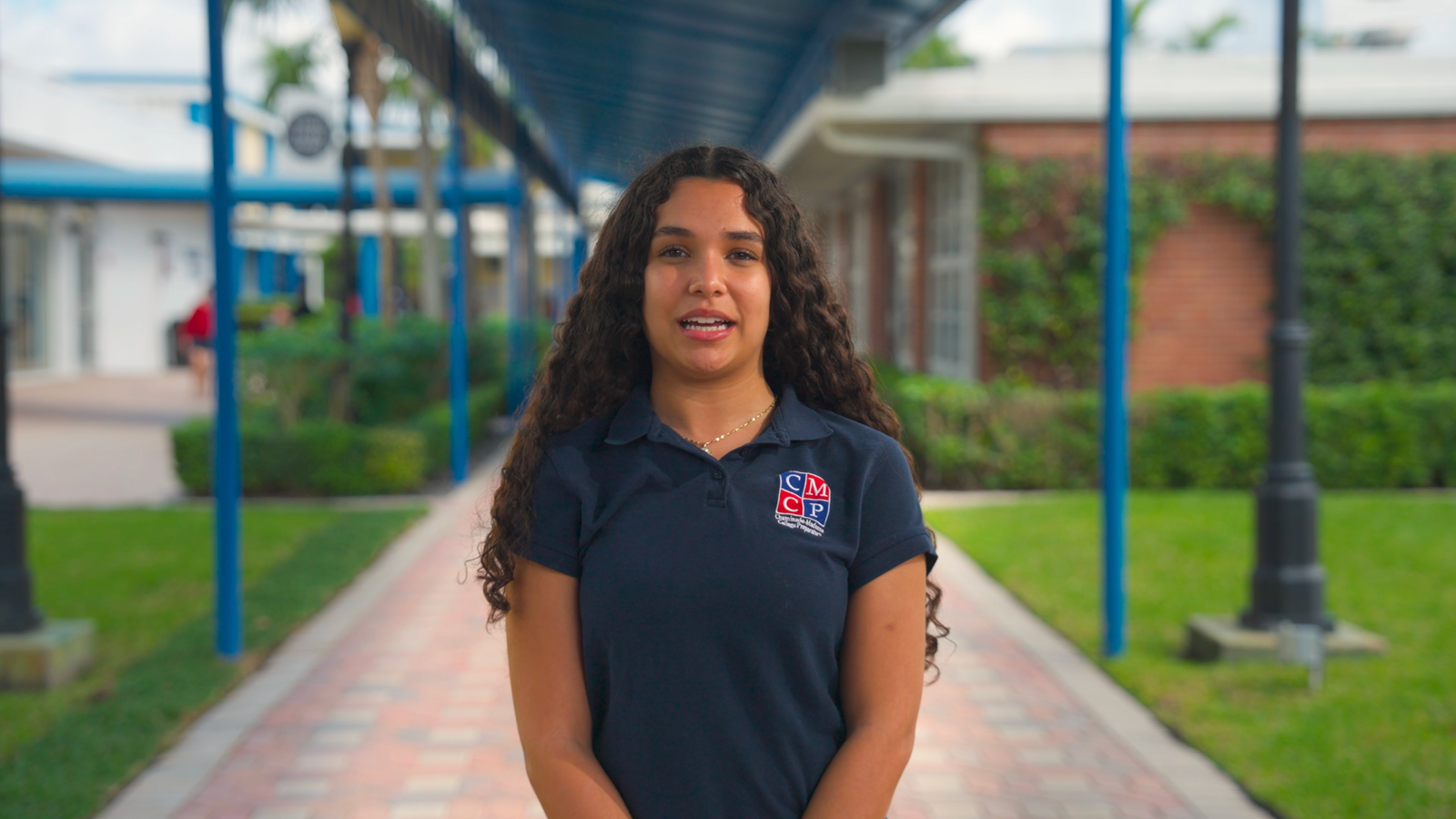 Young woman with long curly hair in a navy polo speaking on a paved walkway